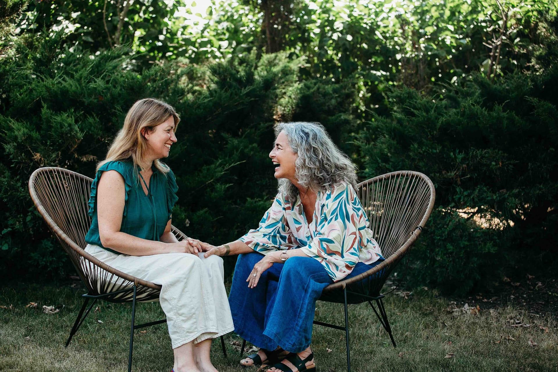 Heidi Feinstein in conversation with a client, the two of them seated in chairs in a garden, hands joined, laughing.