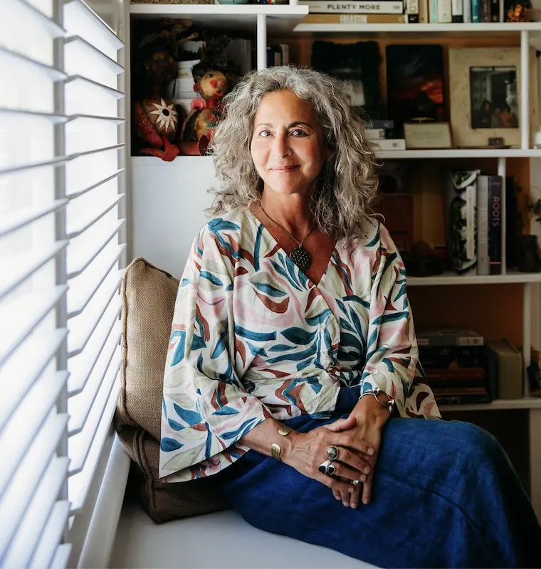 A woman with curly gray hair, smiling at the camera, sitting on a white surface in a room with a bookshelf and window blinds visible in the background.